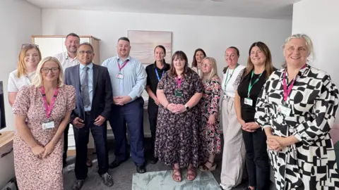 Home Group Twelve members of staff who form the Hope Haven partnership. They are standing in the living room of a new flat. A landscape painting hangs on the wall; a TV and cupboard are also visible in the room.