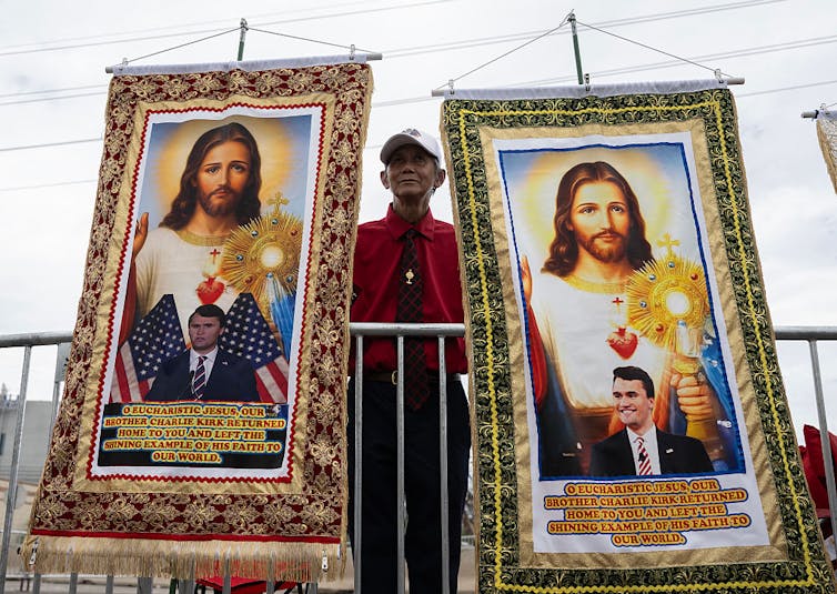 Older man wearing white hate and red dress shirt holds two banners featuring the image of a young man in a suit superimposed over Jesus Christ.