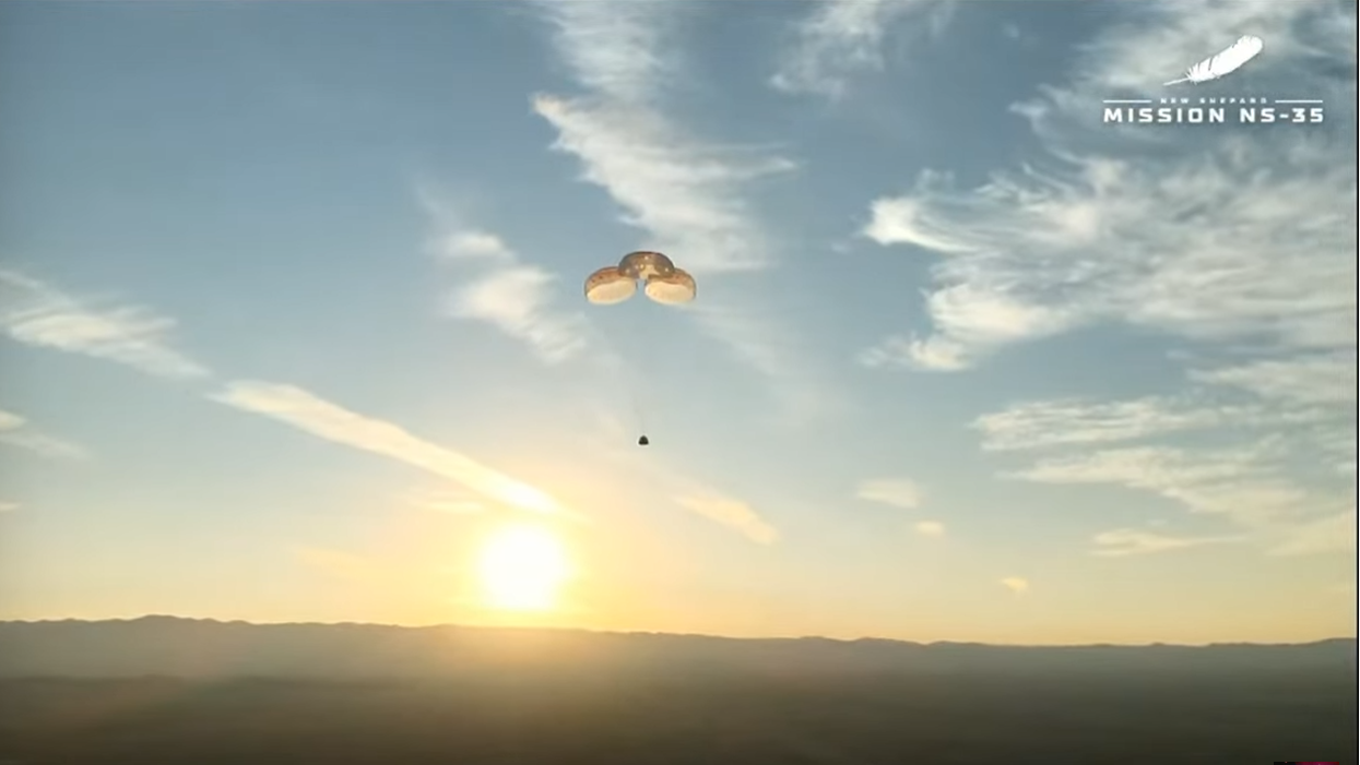 the small silhouette of a space capsule descends under three parachutes in front of the sun.