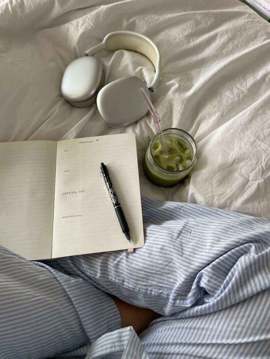 A top-view image of a woman's legs in blue and white stripes pyjamas, Apple headphones, an open journal and black pen and matcha in a glass with a straw.