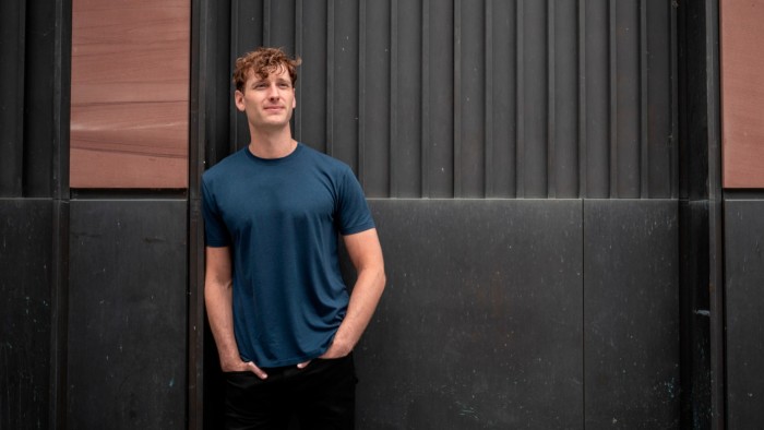 Winston Weinberg stands with hands in pockets against a dark paneled wall, looking slightly upward and smiling.