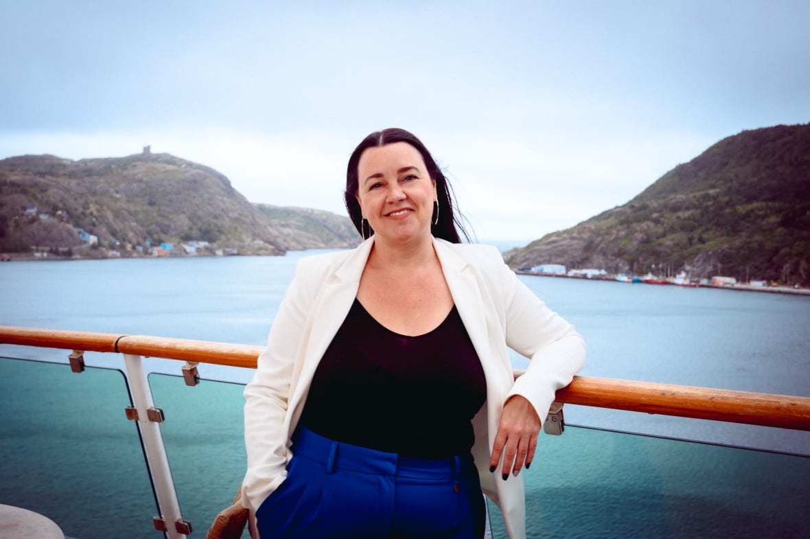 A woman in a white blazer, black t-shirt and blue pants leans against the rail of a cruise ship, with the ocean in the background.