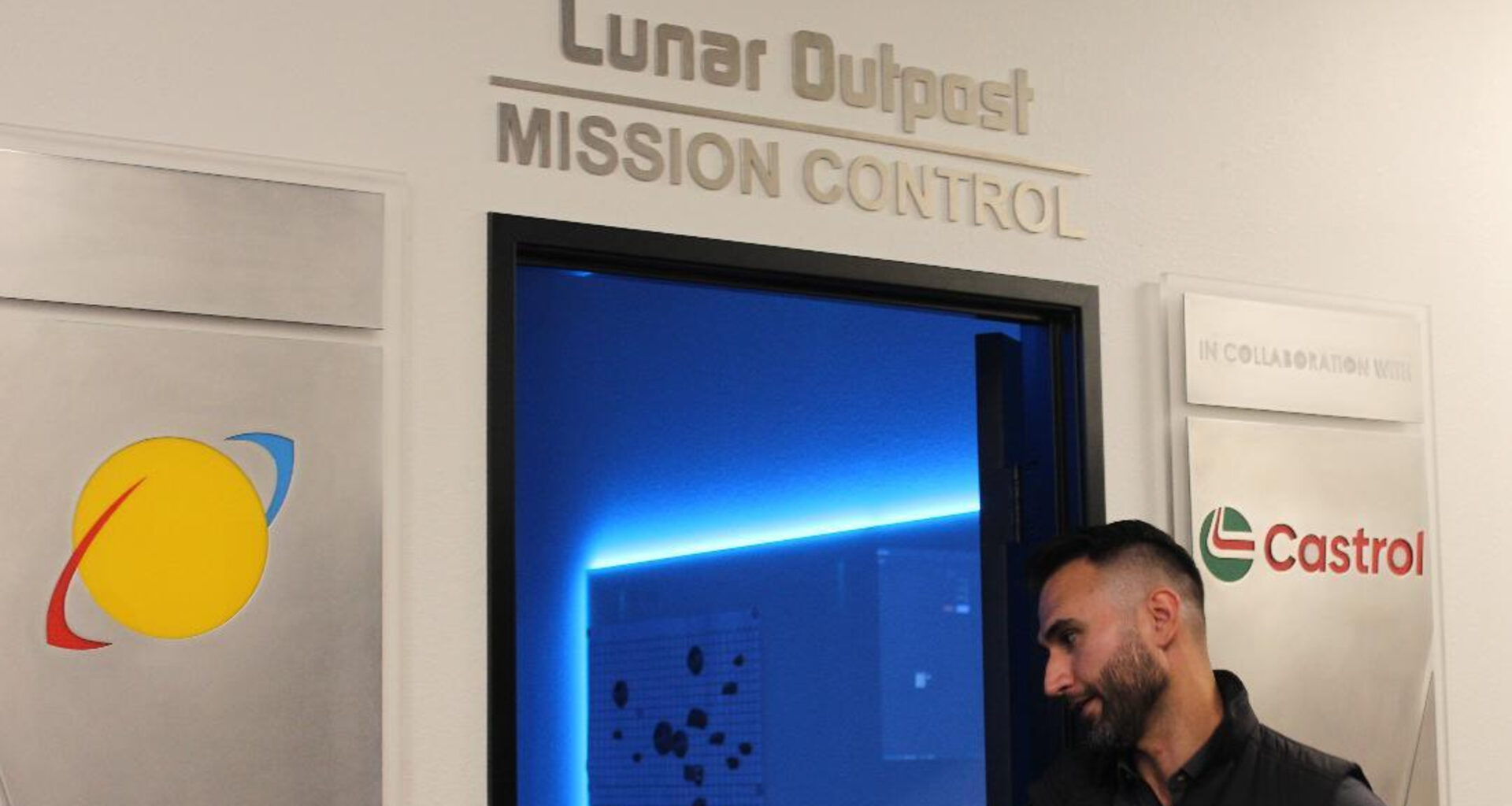 A man wearing a dark polo button up shirt heads into a blue-lit door with the words "Lunar Outpost: Mission Control" above it.