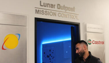 A man wearing a dark polo button up shirt heads into a blue-lit door with the words "Lunar Outpost: Mission Control" above it.