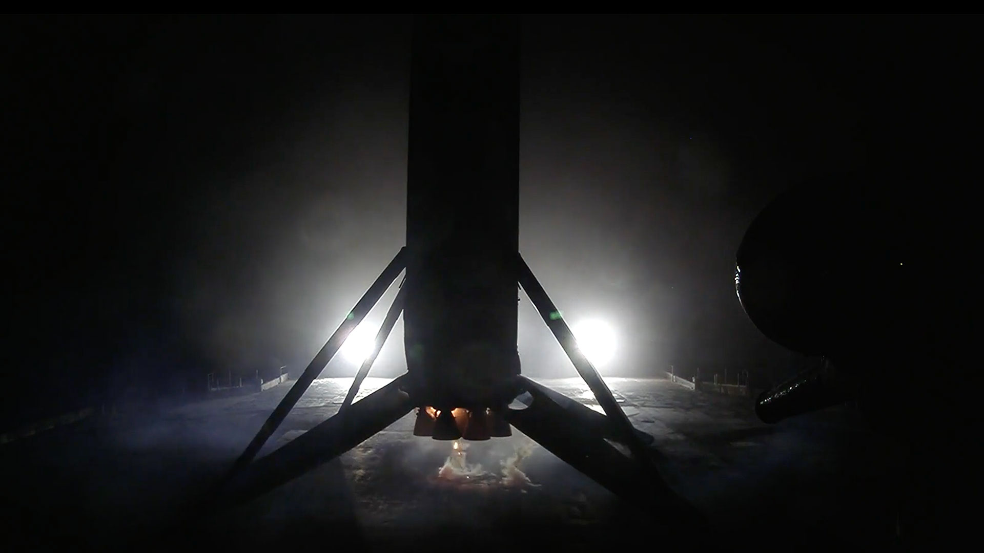the first stage of a rocket is seen in silhouette, backlit by the lights on a sea-based droneship