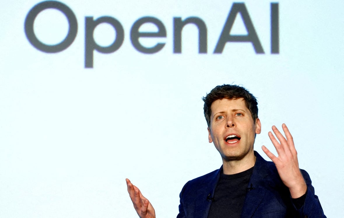 A man gestures while speaking. A large screen behind him says 'OpenAI'