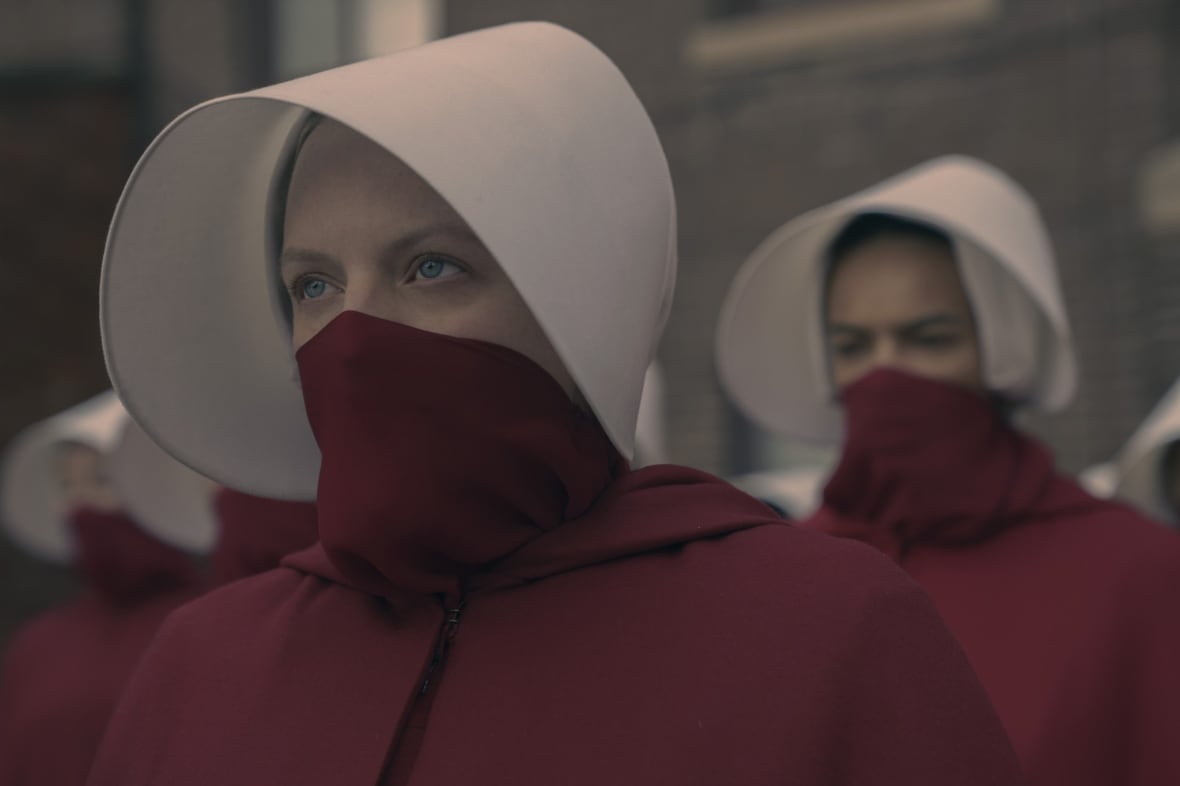 A woman in a red rob, with a red face covering, and a white bonnet on her head stares into the distance while standing in front of other women in similar uniforms. 