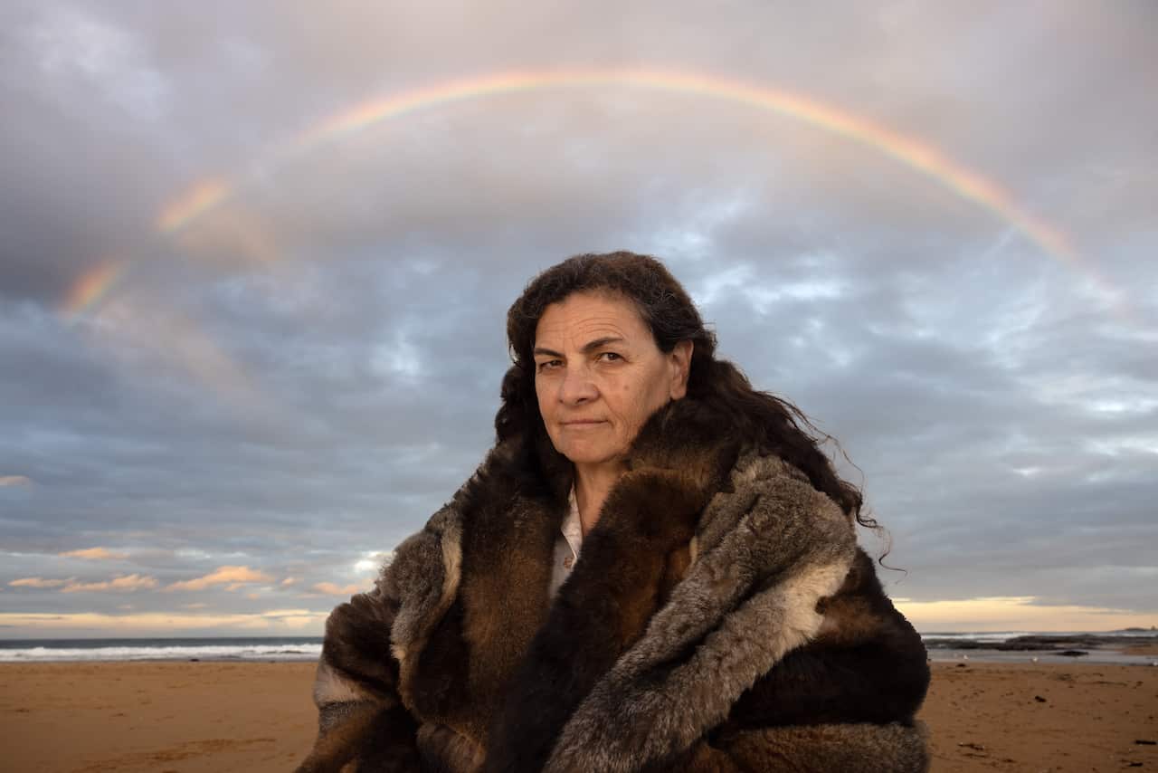 Dr Jodi Edwards, a Walabnja woman in her 50s, wearing traditional possum skins and standing on a shoreline.