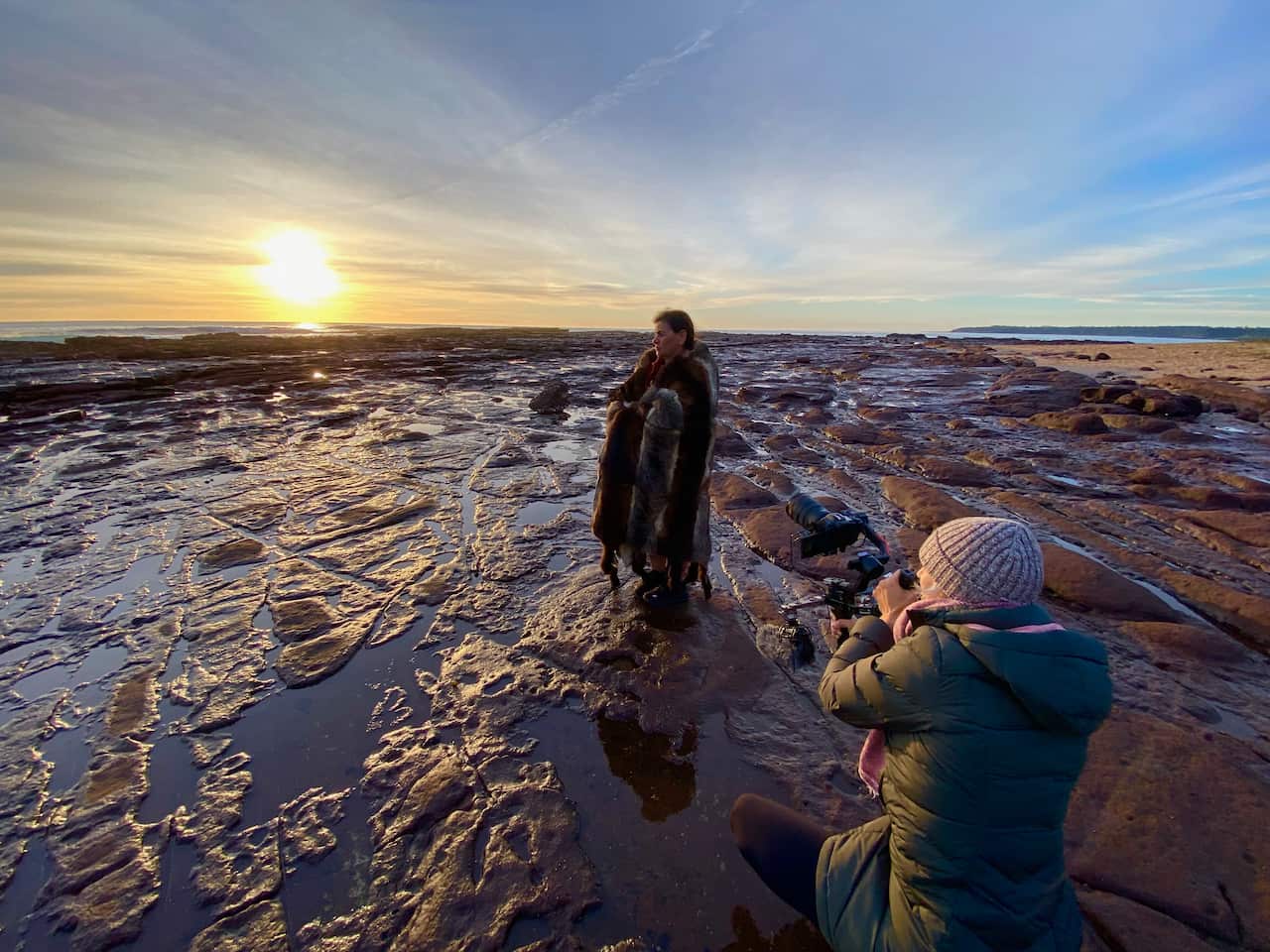 An Indigenous woman wearing a traditional possum skin is seen standing on water-covered rocks, near the ocean at dusk. Another woman is crouched down in the foreground, taking her photo.