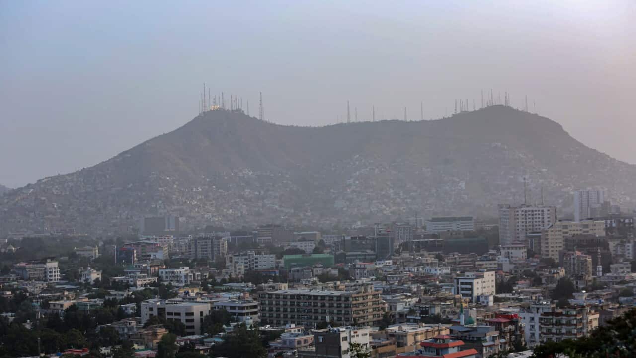 A cluster of houses and buildings with mountains and telecommunications towers in the backdrop.
