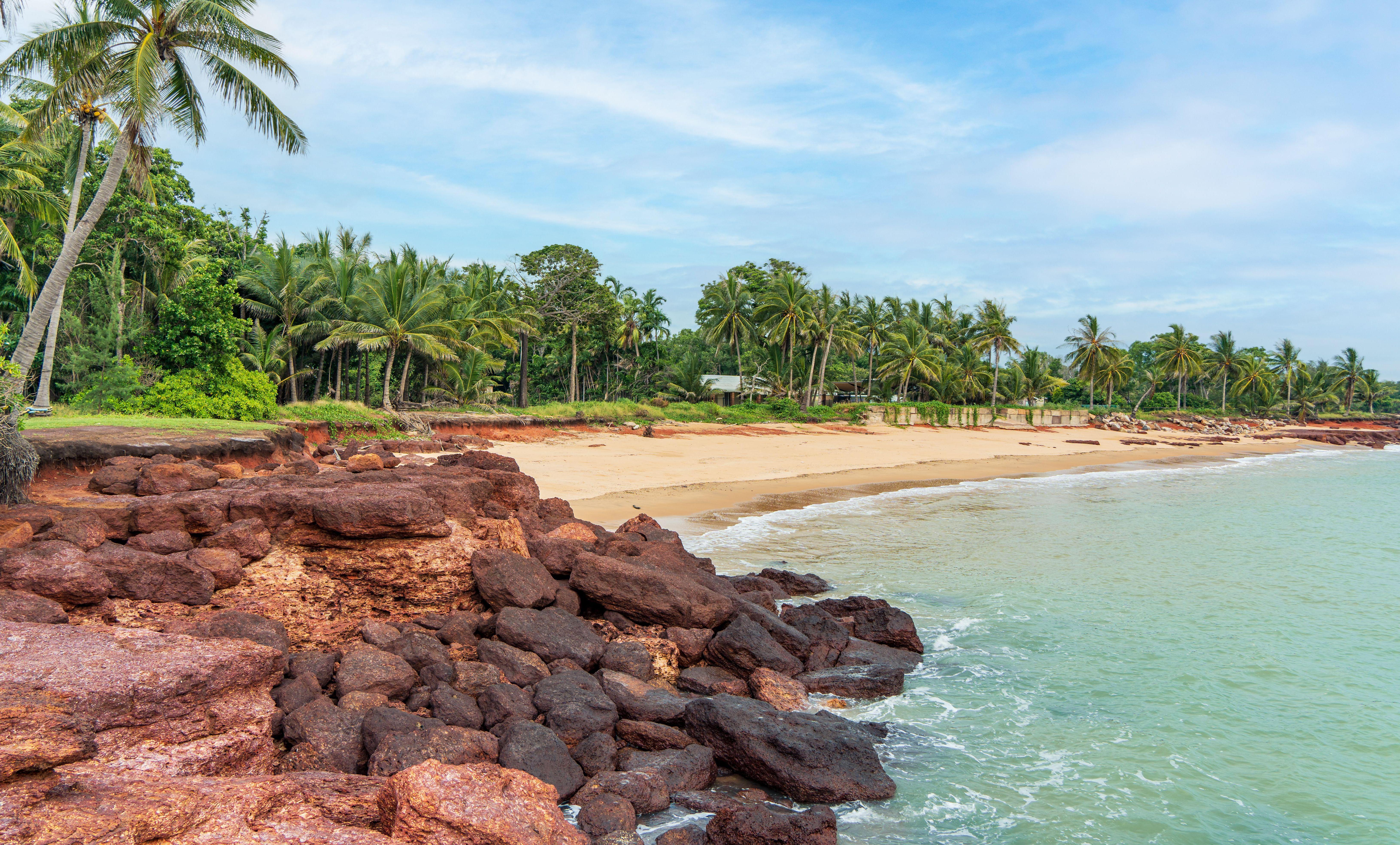 Dundee Beach in the Northern Territory, Australia, featuring a sandy beach, palm trees, and reddish rocks along the coast.