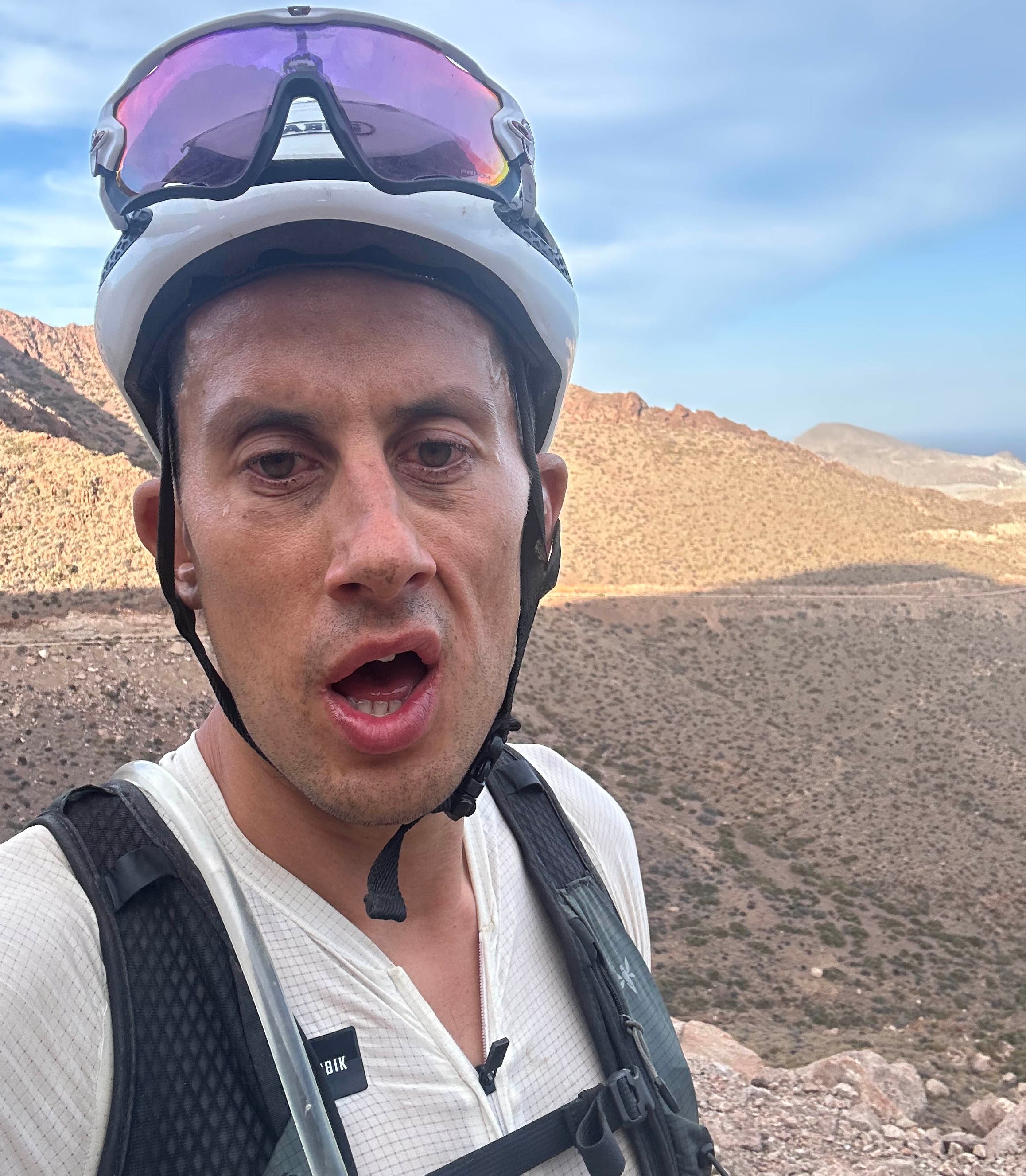 A male cyclist in a white helmet and sunglasses takes a selfie in the desert mountains of Granada, Spain.