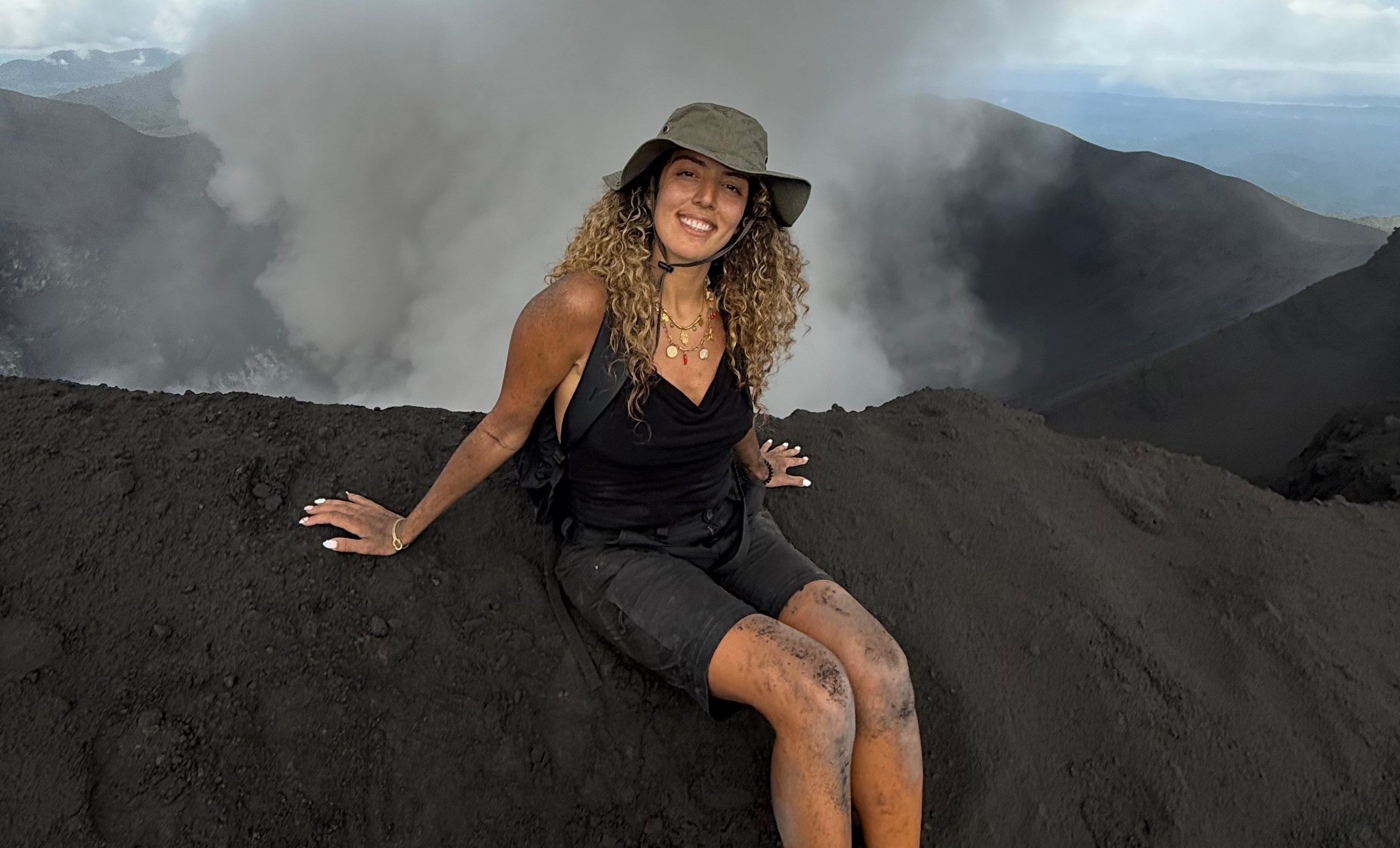 Chaimae Lebyed, a travel influencer, sits on black volcanic ash with smoke plumes rising behind her.