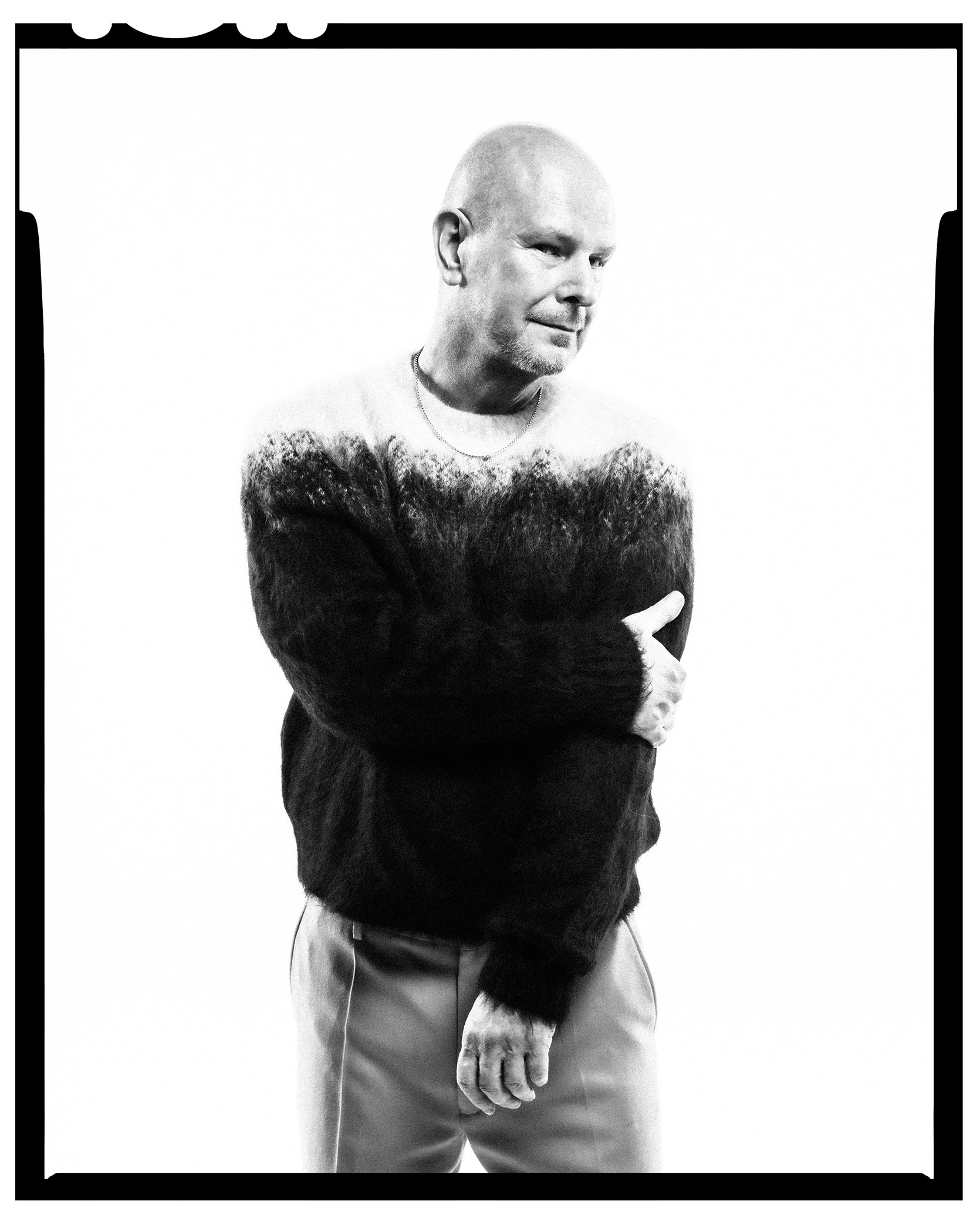 Black and white photograph of a man in a sweater with a light-colored top half and a dark bottom half, his arms crossed, looking to the side.