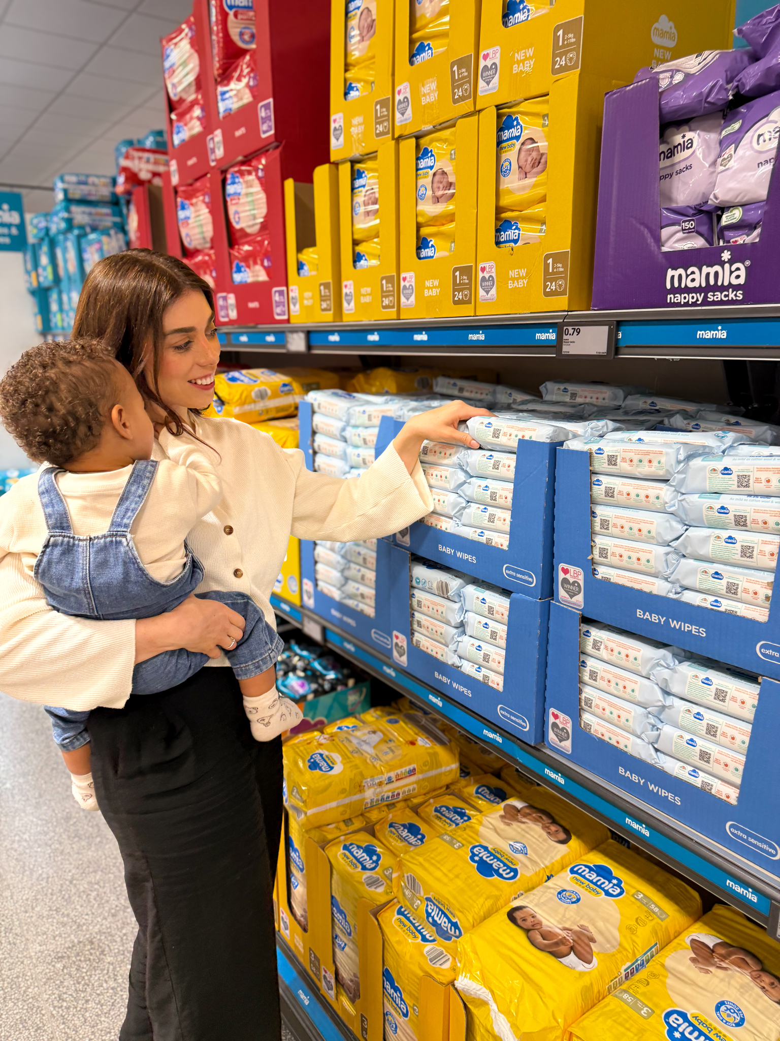 A woman holding a baby while shopping for baby wipes in a supermarket.