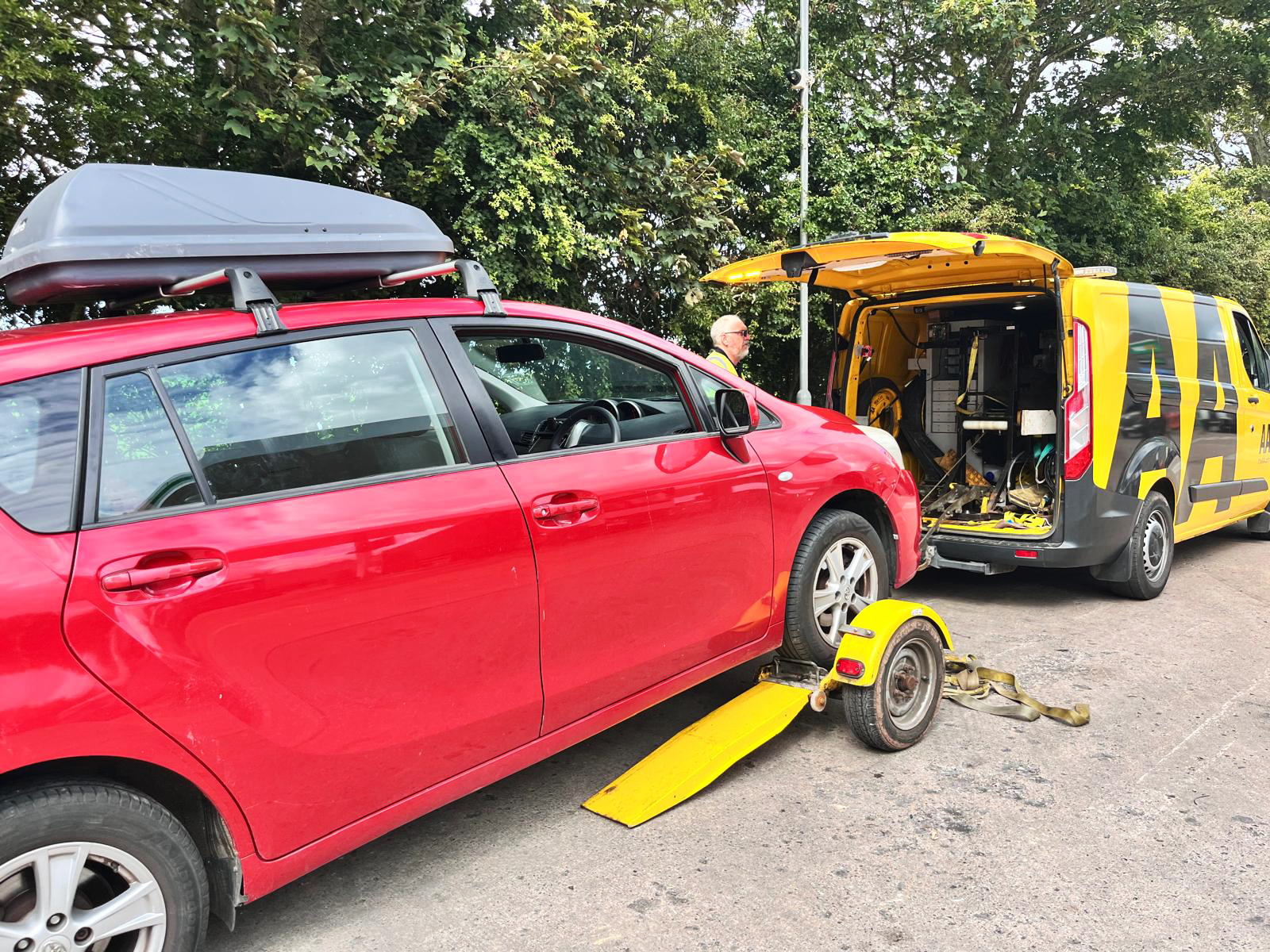 A red car with a roof cargo box being towed by a yellow AA van with its back doors open.