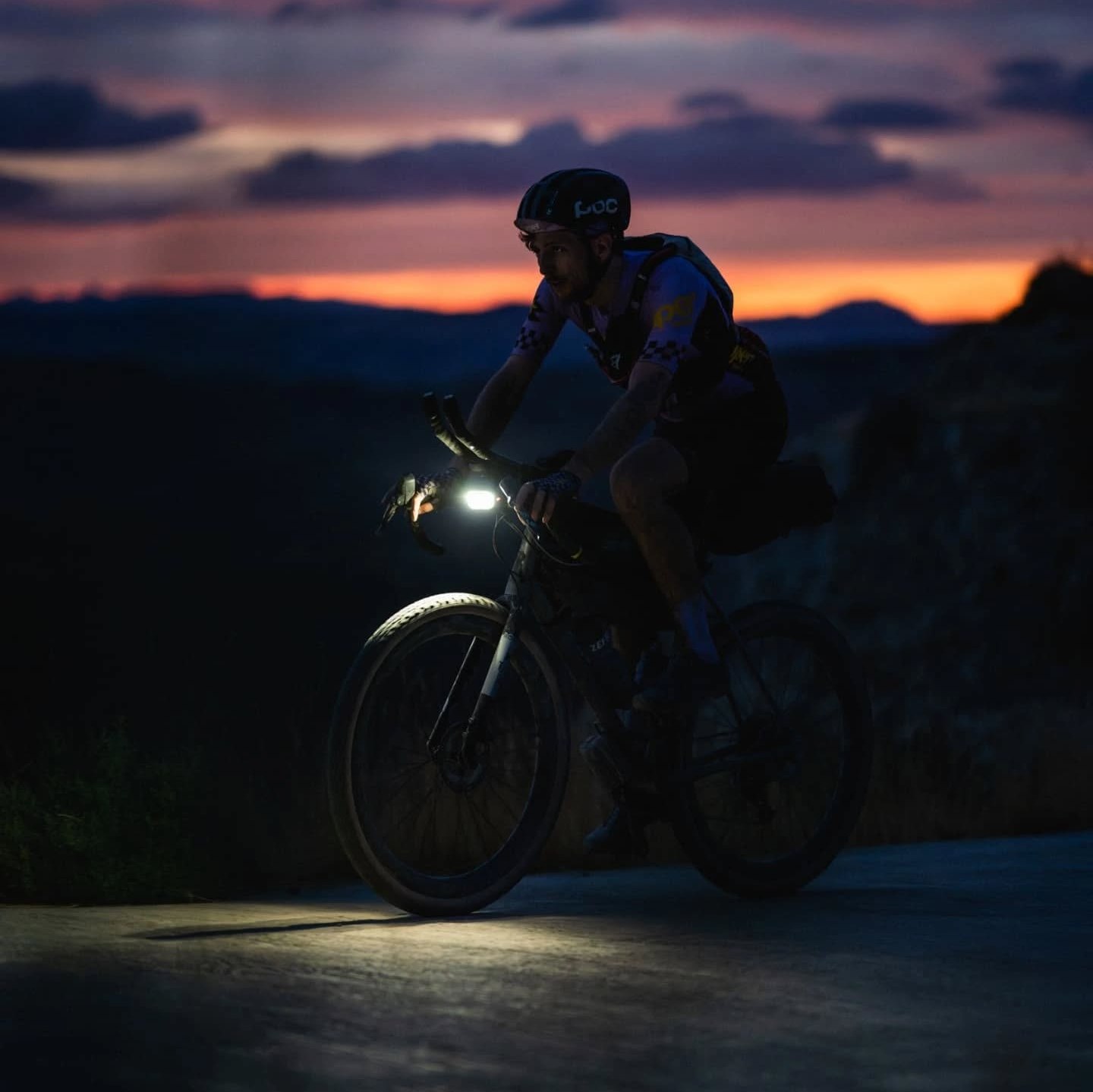 A cyclist riding a gravel bike at dusk, headlamp illuminating the road, with a colorful sky in the background.