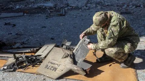 Reuters A police officer in army fatigues crouches, inspecting the remains of what is believed to be a Shahed drone. The floor is entirely rubble