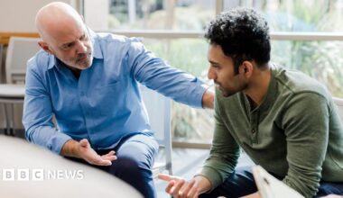 A man in a blue shirt is putting his hand on the shoulder of another man, who wears a green long sleeve top.