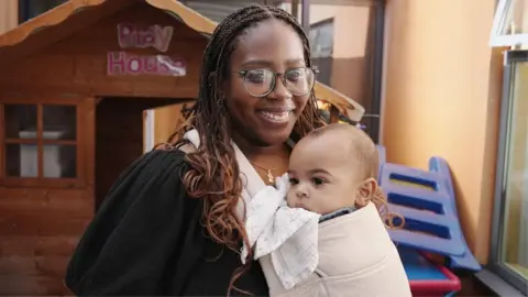 A portrait photo of Jay, a young woman in her late 20s with large round glasses and dreadlocks, and her son Ezra who is being carried in a baby sling. Jay is smiling and looking directly at the camera and Ezra is looking off to one side and chewing on a small white towel.