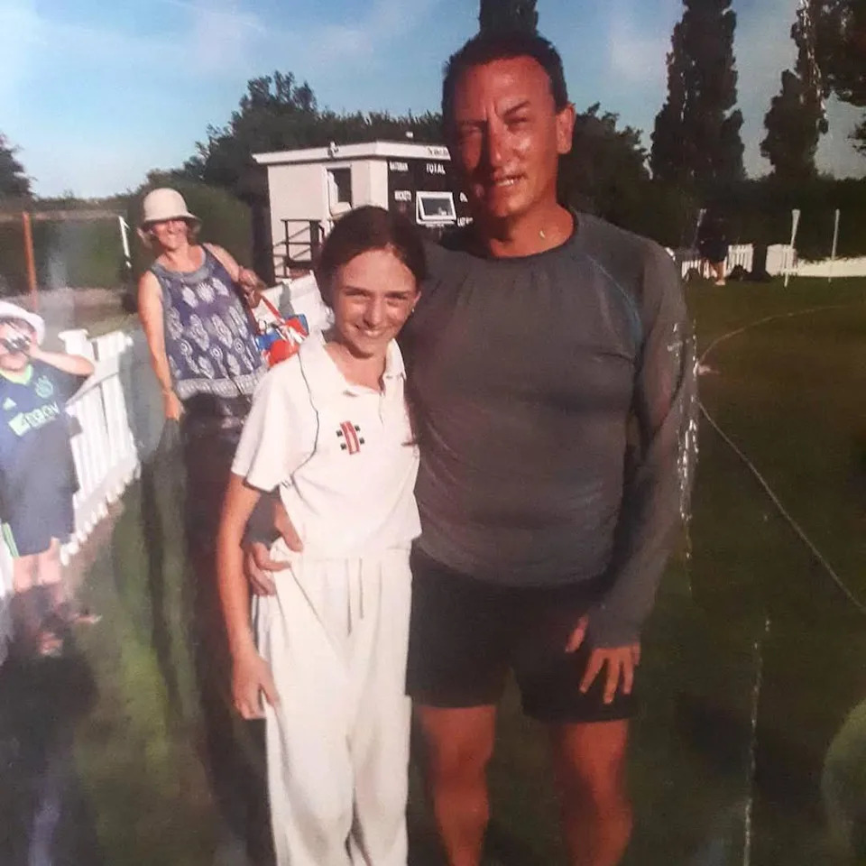 Shortly after representing Wales, Libby is hugged around the waist by her father Michael. Libby is in a white kit with " Cricket Wales / Criced Cymru" logo. A tanned Michael is beaming.