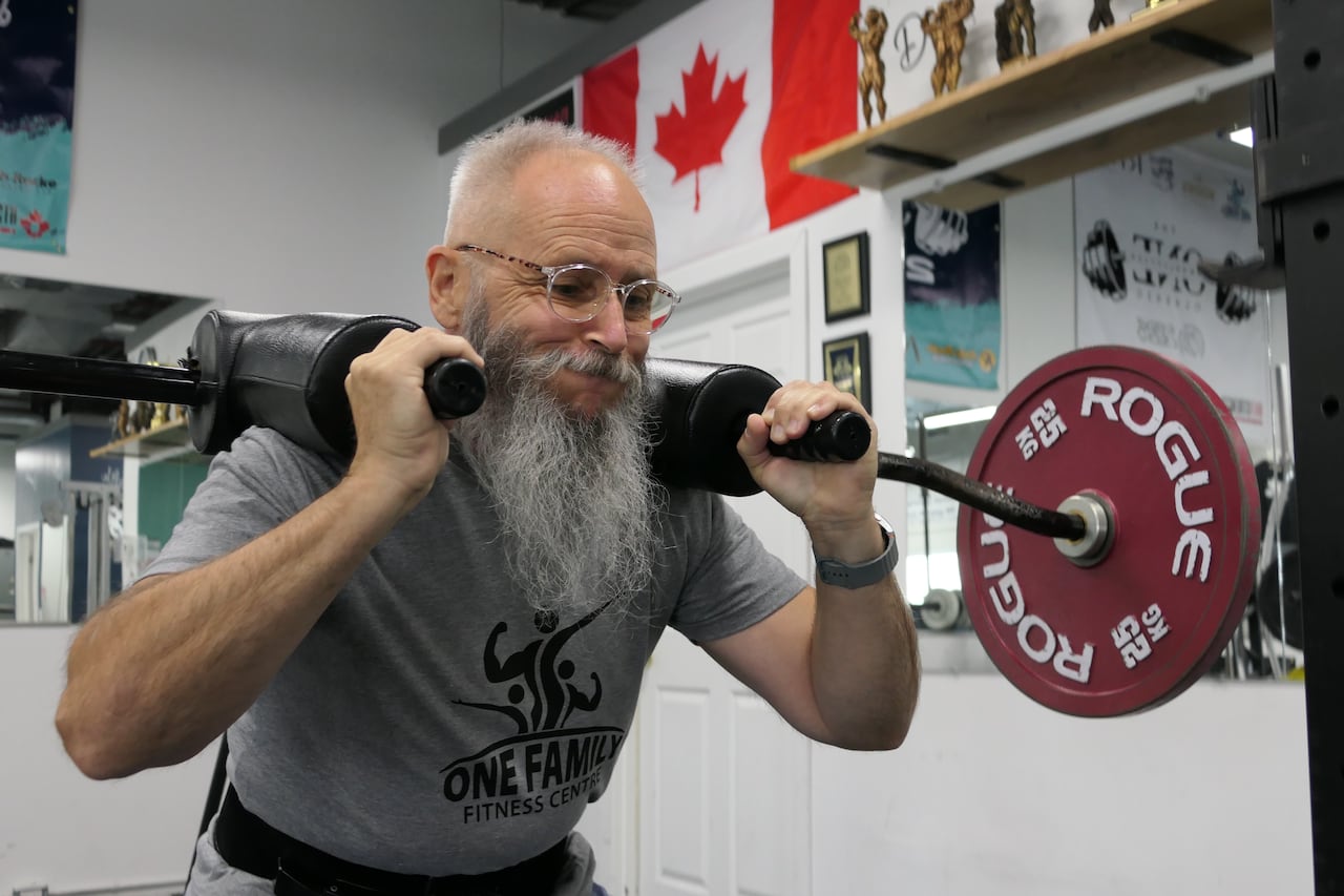 A man in a grey T-shirt, glasses and a big, grey beard squats with weights over his shoulders. His cheeks are puffed. 