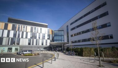 general view of the main entrance to the Royal Hospital for Children and Young People Edinburgh, a modern hospital building with sections of dark grey and light grey panelling and windows. There is a bike rack at the front and a road curves by the entrance