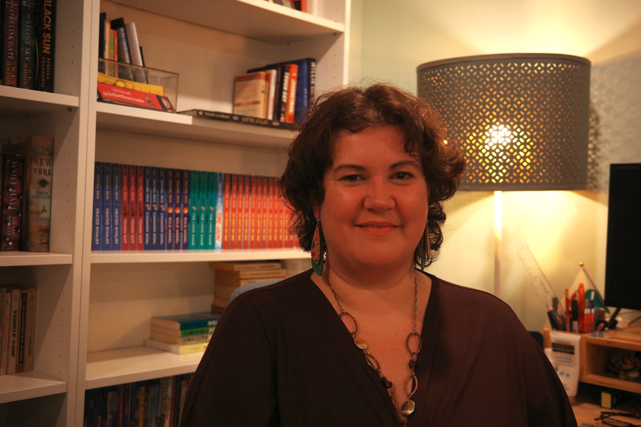 A woman wearing a purple shirt with curly hear sits down in front of a bookcase. 
