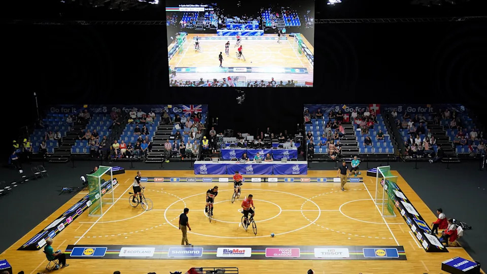 A two-a-side game of cycle-ball takes place on an indoor court with a panel of referees and a crowd behind the far side of the court