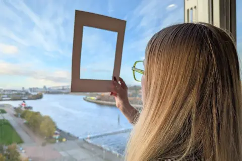 A woman with long, blonde hair looking out a large window at a scenic waterside view. She is holding a rectangular, cream-colored frame up to the window, as if framing a specific portion of the landscape. 