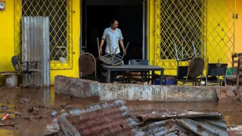 EPA/Shutterstock A man stands in the doorway of a bright yellow building, in front of him the ground is completely  covered in mud with twisted corrugated metal, broken furniture and scattered rubbish. Tables and chairs are also piled up and splattered in mud.