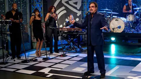 Getty Images Tom Jones sings on stage with a five piece band including backing singers with percussion instruments, a drummer and a keyboard player. He is wearing a navy blue suit, aviator sunglasses with pink lenses and his hair and goatee beard are dyed brown.