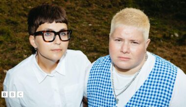 a mid-length image of the band arxx standing in a field. clara is on the left, and has short brunette hair wearing thick-rimmed tortoiseshell glasses, a silver chain and a white shirt. hanni is on the right. they have short platinum blonde hair, and are wearing a silver chain, a white t-shirt and a blue gingham waistcoat
