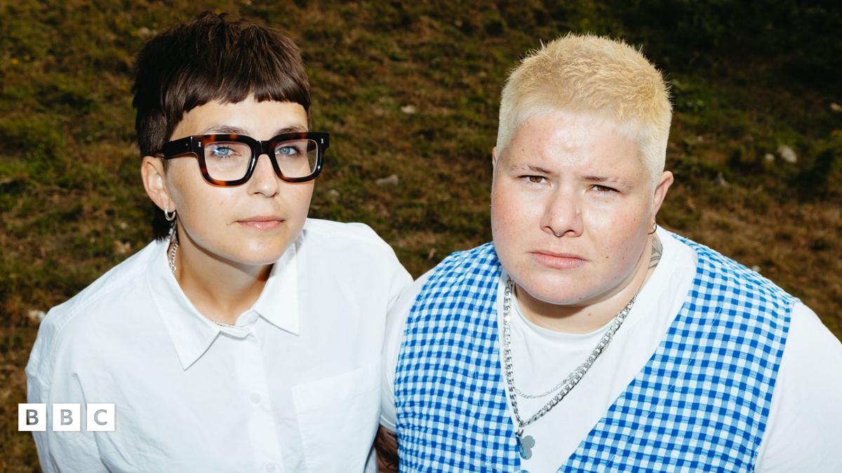 a mid-length image of the band arxx standing in a field. clara is on the left, and has short brunette hair wearing thick-rimmed tortoiseshell glasses, a silver chain and a white shirt. hanni is on the right. they have short platinum blonde hair, and are wearing a silver chain, a white t-shirt and a blue gingham waistcoat