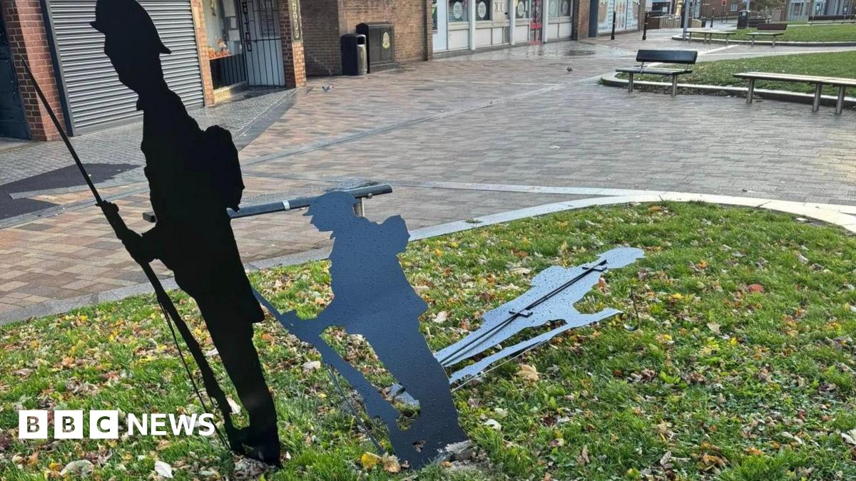 Three black metal silhouette sculptures depicting soldiers. One figure holding a rifle with a bayonet is standing upright while the second figure is bent lower and the third one near the ground. They are placed on a small grassy area with scattered autumn leaves, surrounded by a paved pedestrian space.