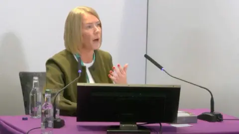 Lampard Inquiry / YouTube Laura Cozens gives evidence to the Lampard Inquiry. She sits at a purple table with two microphones and a computer monitor in front of her. She has shoulder-length blonde hair and wears a green jacket over a white t-shirt.