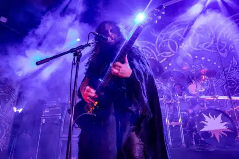 Getty Images Nathan Weaver from Wolves in the Throne Room performs on stage with a purple backdrop. He has long hair and is singing into a microphone also playing a guitar. 