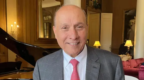 Tony Fisher/BBC A man wearing a suit and a red tie standing in front of a grand piano in a large room with sofas and paintings on the wall.