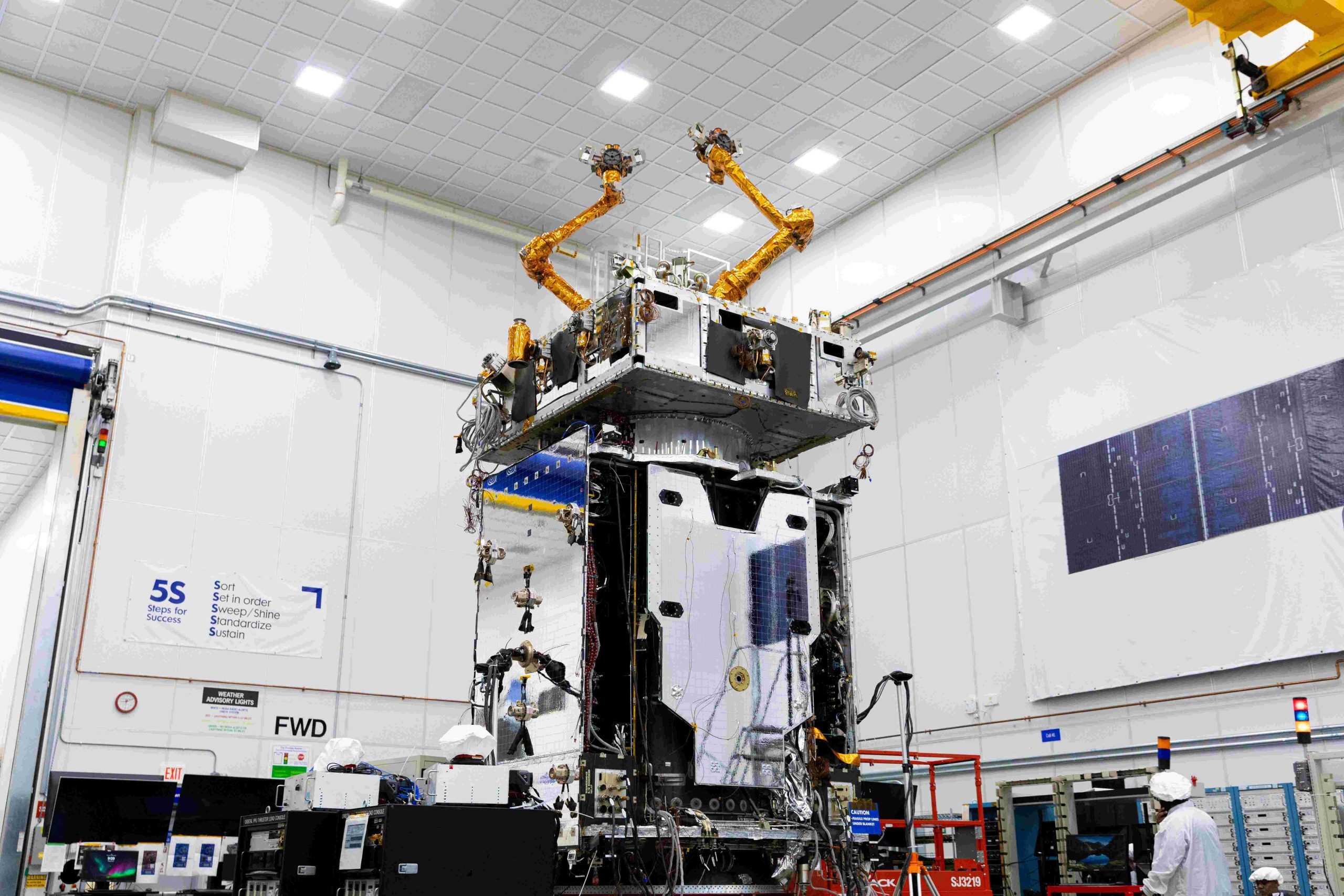 Inside a cleanroom in a warehouse, a satellite is worked on using various robotic tools