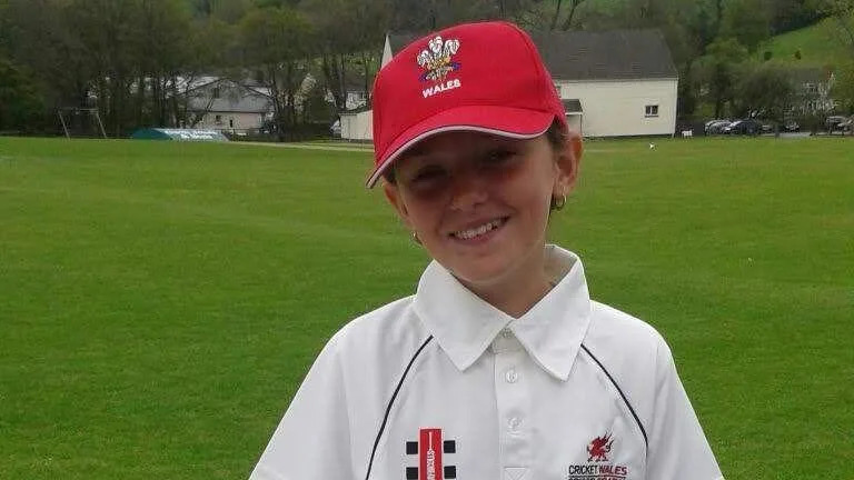 Libby wearing her white "Welsh Cricket/Criced Cymru" polo shirt and long white trousers and a red Wales cap, pictured as a young teenager. She is standing in front of a white picket fence at a rural-looking cricket ground