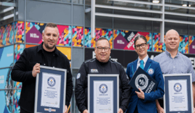Three men and two women stand side by side in front of a large, modern building with colourful designs on the exterior. Each person is holding a framed certificate from Guinness World Records, which features a blue and white design with the official logo at the top. They are facing the camera. The award reads: "The most users in a mental health lesson was achieved by Think Mental Health, Think Cloud, Hull College, Smith & Nephew (all UK) in Hull, UK, on 10 October 2025."