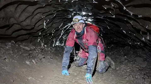 Handout A man wearing protective outdoor clothing and a helmet with a torch on his hands and knees inside a tight, rocky space.