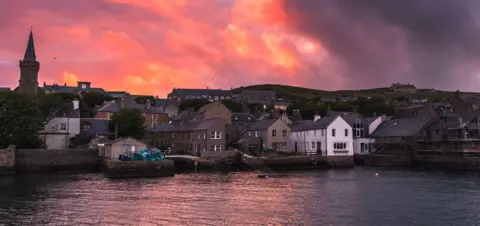 Getty Images A cluster of buildings along a waterfront. The buildings are varied in colour and architecture, suggesting a historical development over time. In the background, a hillside dotted with more buildings is visible. The sky is a dramatic mix of pink, orange and purple hues.
