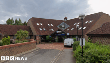 A brick building with a large sloped tile roof. There is a Blue sign over the entranceway that reads Priority House.