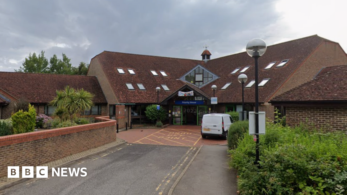 A brick building with a large sloped tile roof. There is a Blue sign over the entranceway that reads Priority House.