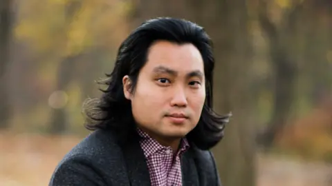 Es Lee Wearing a grey blazer and checked shirt Es Lee looks into the camera.