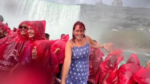 Holly Hyde Holly Hyde standing at Niagara Falls - she is wearing a light blue dress with white flowers dotted on it. Niagara Falls is visible behind her, and she is surrounded by people in plastic red ponchos - Holly stands out as she is not wearing a poncho and is smiling at the camera while covered in water  