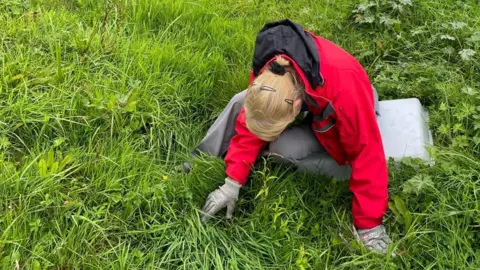 Wiltshire Wildlife Trust A woman sits on a plastic box in amongst lush grass. She is looking downwards at the grass with gloved hands, looking through the blades. She wears a red raincoat and grey trousers.