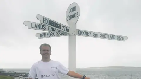Drew Jonksman Drew Jonkmans is pictured dressed in a white T-shirt. He is a young man with short blonde hair and he is smiling. He is standing in front of a signpost marked John O' Groats. The sign has fingers pointing to Edinburgh, Land's End, New York, Orkney and Shetland. The sea can be seen in the background.
