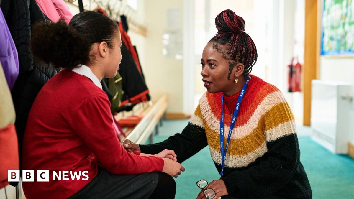 The image shows a woman and a girl in a school cloakroom, with the little girl sat on a bench while the woman crouches in front of her. The woman, a teacher, is wearing a striped jumper while holding a pair of glasses with black frames. The little girl is wearing a red jumper and a grey skirt with black tights. The floor is green and coats of different colours can be seen in the background.